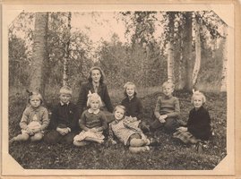 Svart vitt foto, sju skolbarn och lärarinna sitter i naturen